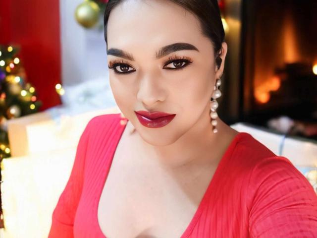 BoobsieBigCock with dark red lipstick, thick eyebrows, and long eyelashes, wearing a red top, and dangling earrings, has a sexy, confident look in a Christmas-themed photo.