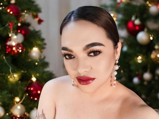 BoobsieBigCock with dark hair, red lipstick, and pearl earrings, looking at the camera with raised eyebrows, standing in front of decorated Christmas trees. Boobs are not visible.