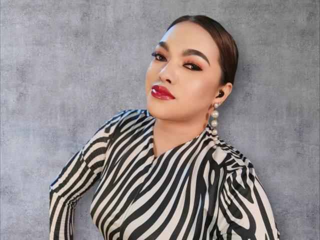BoobsieBigCock has dark brown hair pulled back, wearing a zebra-striped top, red lipstick, and pearl earrings, with a sexy, confident expression. BoobsieBigCock's makeup is bold and elegant.