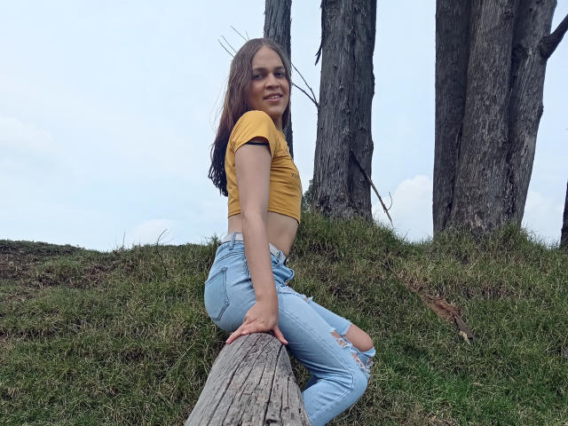 CoulpleDark with brown hair, yellow crop top, and ripped blue jeans, sitting on a wooden log, smiling, outside with tall trees, grassy hill, and a cloudy sky.