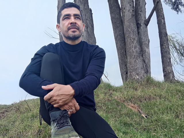 CoulpleDark sits on grass with legs crossed, wearing a black long-sleeve shirt and black pants, looking up with a slight smile, surrounded by tall trees.