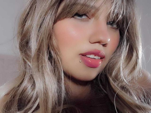 AvririzoX has wavy blonde hair, pink glossy lips, a silver lip piercing, and a shiny nose, looking slightly to the side with a soft, seductive smile.