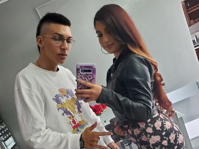 Diamantes69 with red hair in a black leather jacket and floral skirt takes a mirror selfie with Diamantes69 in a white graphic sweatshirt and glasses. Diamantes69 points.