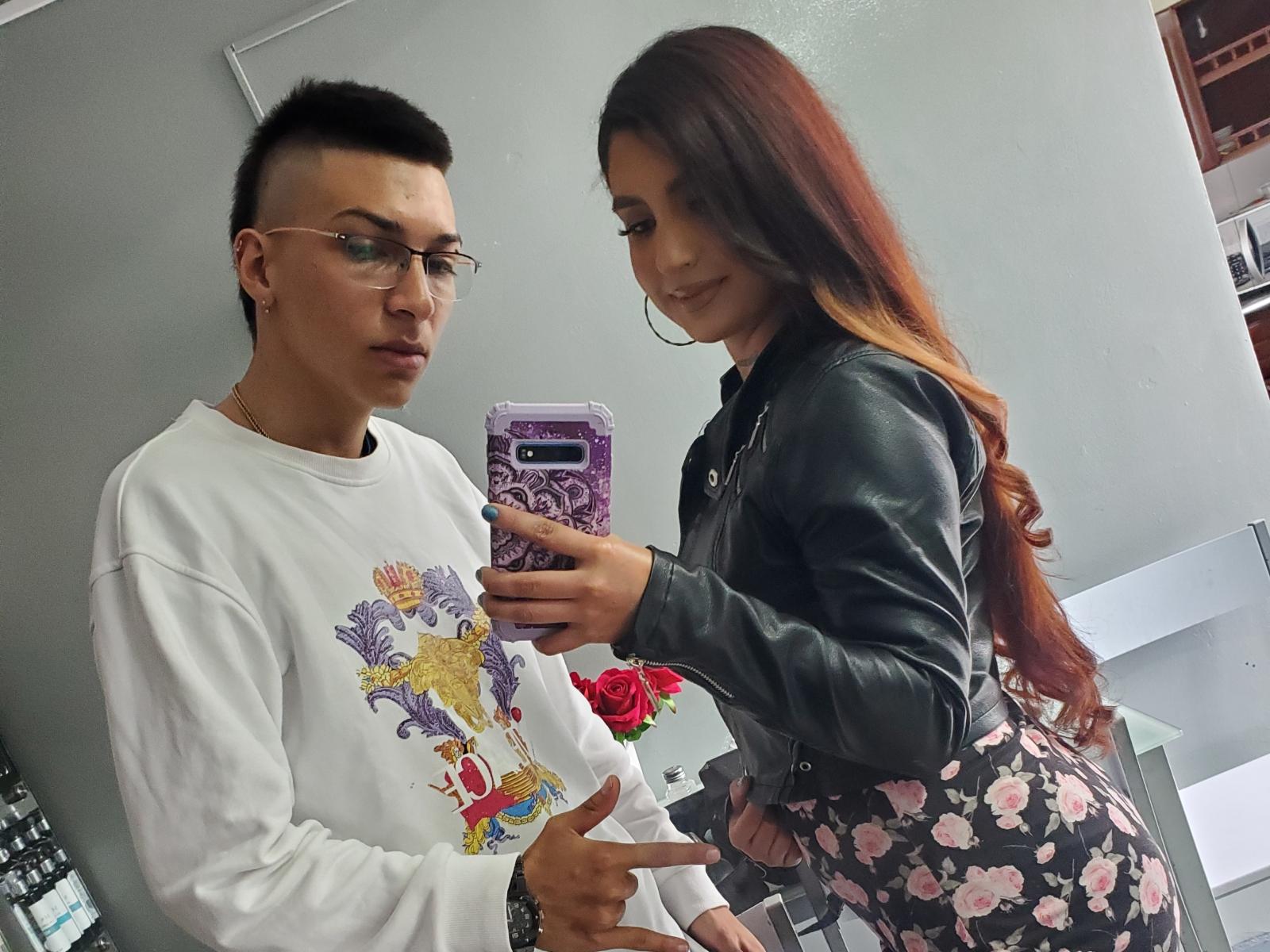 Diamantes69 with red hair in a black leather jacket and floral skirt takes a mirror selfie with Diamantes69 in a white graphic sweatshirt and glasses. Diamantes69 points.