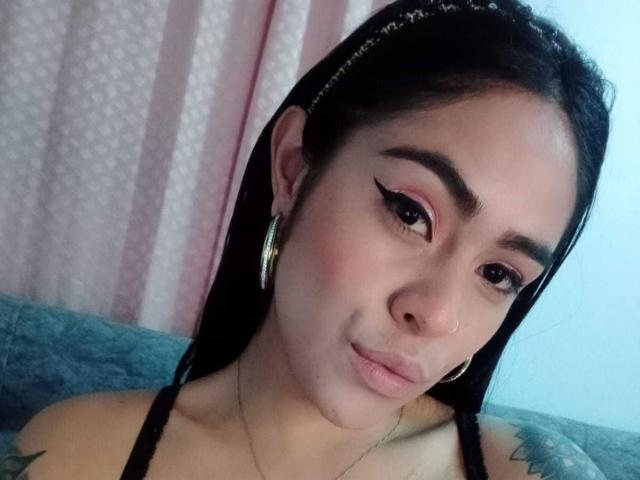 StellaBeckker has medium skin, dark straight hair, bold eyeliner, pink eyeshadow, small nose ring, gold hoop earrings, and subtle makeup, with a slight pout on her face.