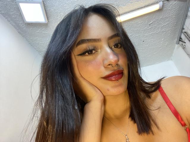 MiraCataleyax with dark brown hair, red lipstick, winged eyeliner, nose ring, and red bra, holding her cheek, smiling, ceiling light and textured ceiling in the background.