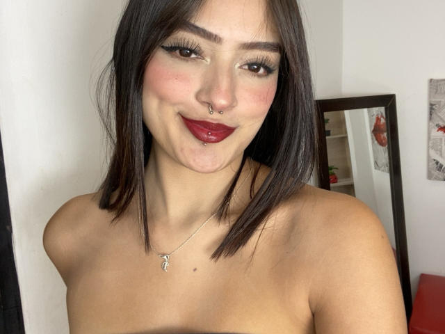 MiraCataleyax has medium brown, straight shoulder-length hair, dark red lipstick, a nose piercing, and a necklace with a small pendant. She's topless, showing her bare shoulders.
