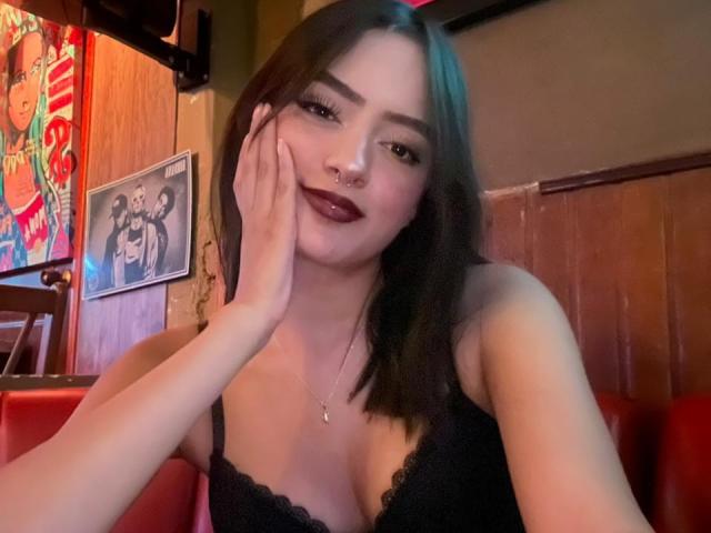 MiraCataleyax with dark hair, nose ring, and dark lipstick, touching her cheek, wearing a black lace top, showing her tits, in a red booth.