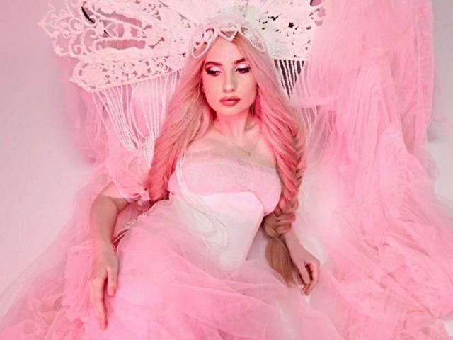 ElzaKomx with pink hair, lace headpiece, and white strapless dress, surrounded by pink tulle, lying down, showing her tits and pink makeup, looking sexy.