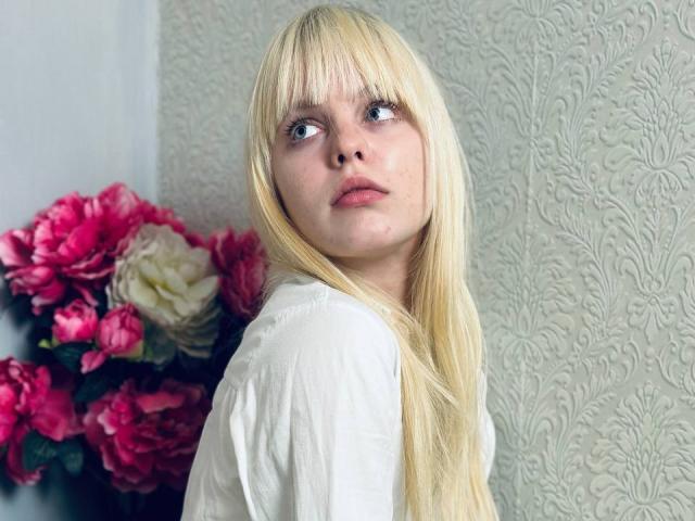 Lialit has long blonde hair, bangs, and is wearing a white shirt, looking up with a slightly confused expression. Pink and white flowers are in the background.