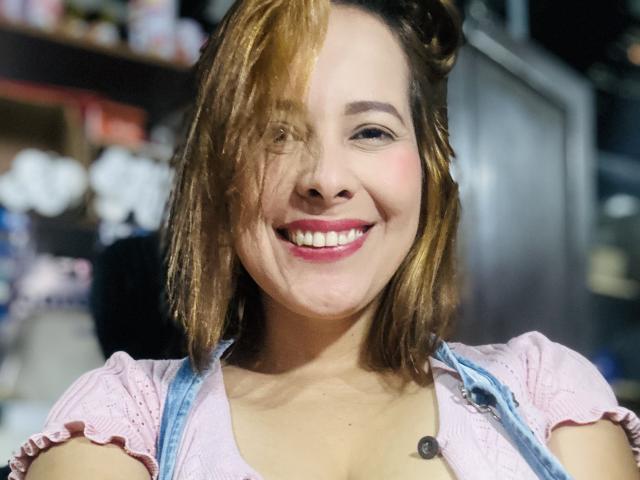 SaritaVelez with dirty blonde hair, smiling, pink puffed-sleeve top, button visible, blue denim straps, pink lipstick, cute, tits slightly visible, medium skin, small black nose tattoo.