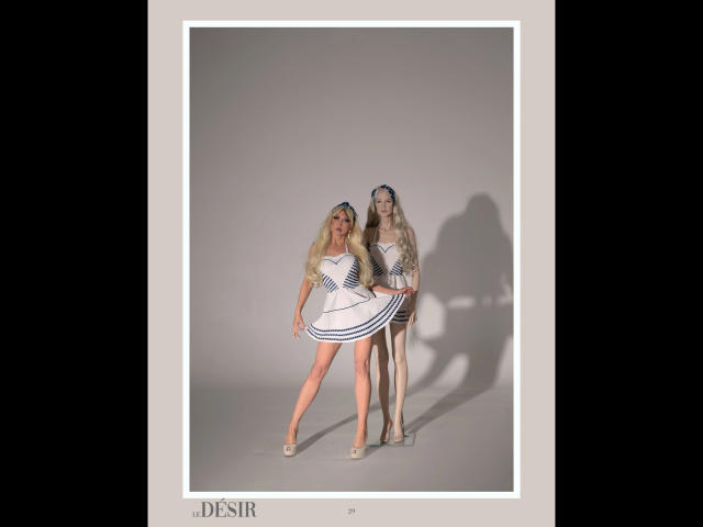 Two Shezel-hots with long blonde hair, wearing white and blue striped dresses, standing and smiling, shadows on the wall, in high heels, on a gray background.
