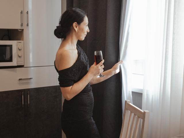 LiluGrants stands in a kitchen, holding a glass of red wine, wearing an off-shoulder black dress, looking out a window with white curtains. Her ass is visible.