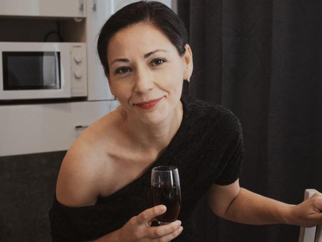 LiluGrants with black hair, red lipstick, and bare shoulder, smiling, holding a glass of red wine, wearing a black off-shoulder top.