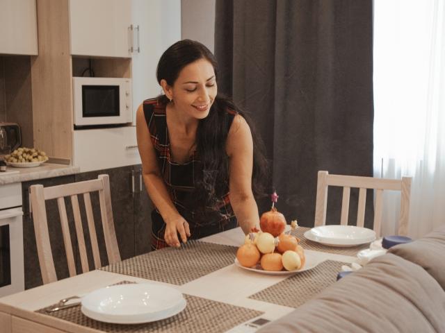 LiluGrants, dark-haired, wearing a plaid apron, bends over a table, smiling, touching a plate of apples with a pink candle on it. Kitchen, wooden chairs, white plates.