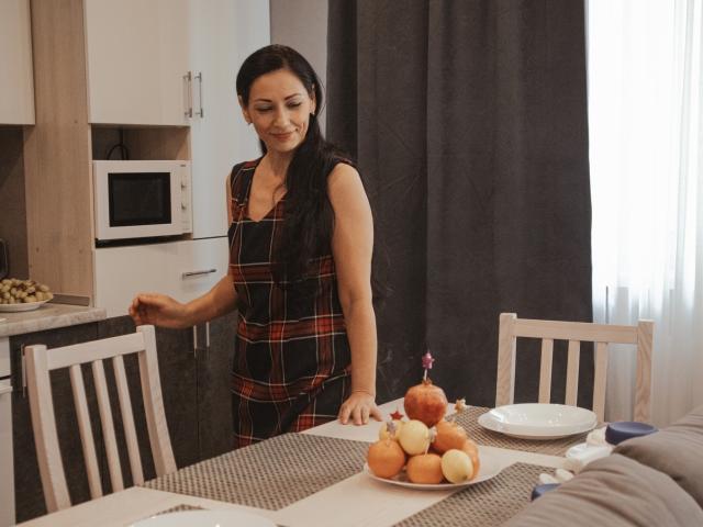 LiluGrants with long black hair, wearing a plaid dress, stands in a kitchen, smiling, touching a counter, next to a table with apples and a pumpkin.