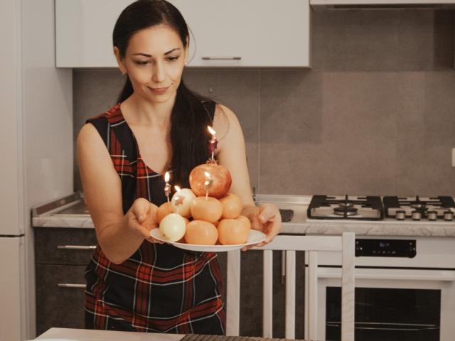 LiluGrants with dark hair, wearing a plaid apron, holds a plate of orange and white eggs with lit candles in a modern kitchen. She's smiling, looking down.