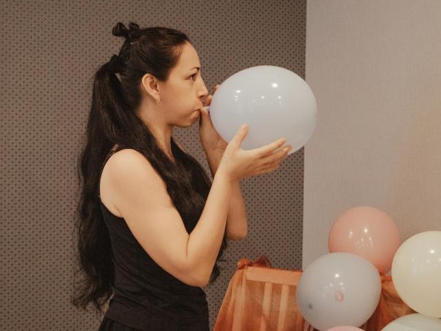 Lilu, met lang zwart haar in een staart, een zwarte tanktop, blaast in een witte ballon, handen in de buurt van haar mond, omringd door roze en witte ballonnen..
