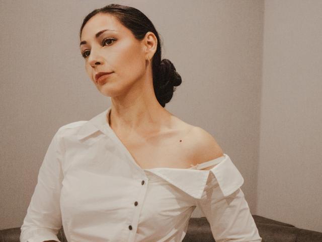 LiluGrants with dark hair in a bun, wearing a white off-shoulder button-up shirt, showing a small mole on her right shoulder, looking to the side.