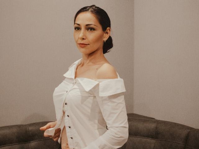 LiluGrants with dark hair, wearing an off-shoulder white button-up shirt, showing her tits, smiling subtly, standing in front of a grey couch.