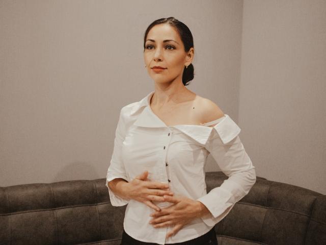 LiluGrants with dark hair, wearing an off-shoulder white shirt, has her right hand on her belly, standing in front of a gray couch, looking confident.
