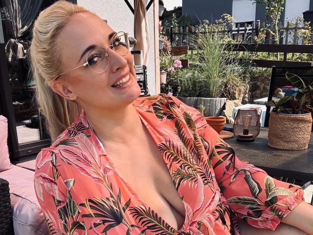 JillFox-hot with blonde hair, glasses, and a pink leaf-patterned shirt, showing cleavage, smiling outdoors, sitting on a wicker chair. Her tits are noticeable.