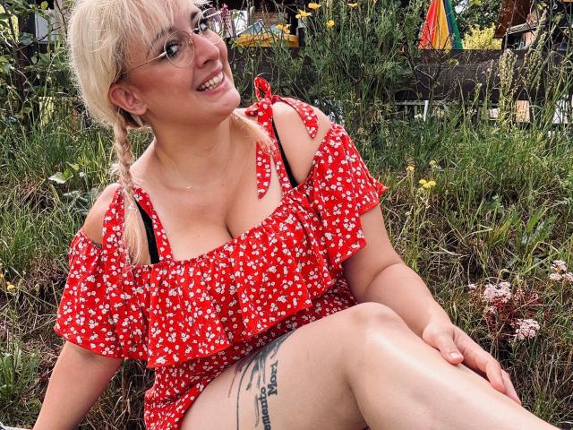JillFox-hot with blonde hair, glasses, red floral dress, exposed tits, black strap, tattoo on thigh, smiling, sitting in grass.