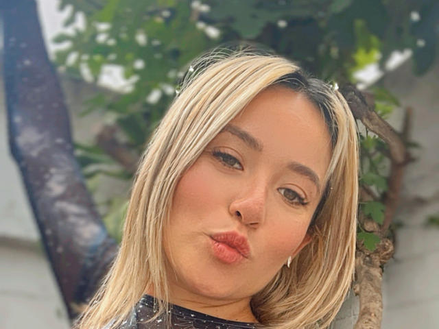 NickySexFire has blonde hair, puffy pink lips, and a cute face with brown eyes, making a pouty expression against a tree with green leaves.