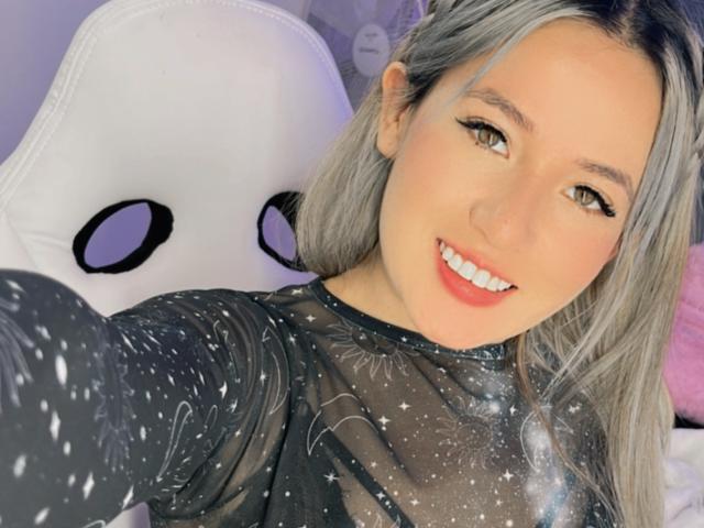NickySexFire with blonde hair, smiling, wearing a black sheer top with star patterns, and a white chair with black eyes in the background. Boobs are not visible.
