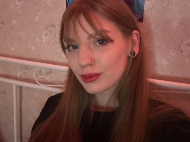 ElsaBj has straight brown hair, blue eyes, red lipstick, black ear gauges, and a nose ring, wearing a black shirt, with a slight smile in a softly lit room.