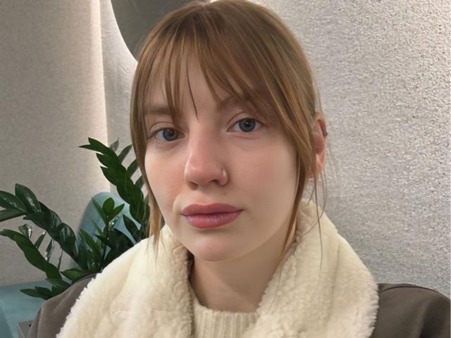 ElsaBj with pale skin, blue eyes, and brown hair in a ponytail, wearing a white, fluffy scarf, has pink lips and a small nose, looking straight at the camera.
