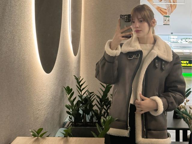 ElsaBj with puffy brown jacket, white fur lining, holding phone, green plants in background, standing in cafe, taking mirror selfie, light brown hair, subtle smile, white knit shirt.