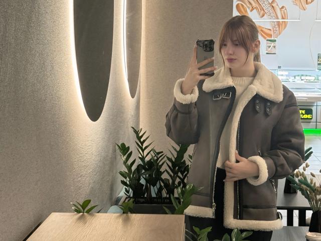 ElsaBj in a brown parka with white fur trim, standing, holding phone, reflection in mirror, green plants in background, light brown wall with oval cutouts.