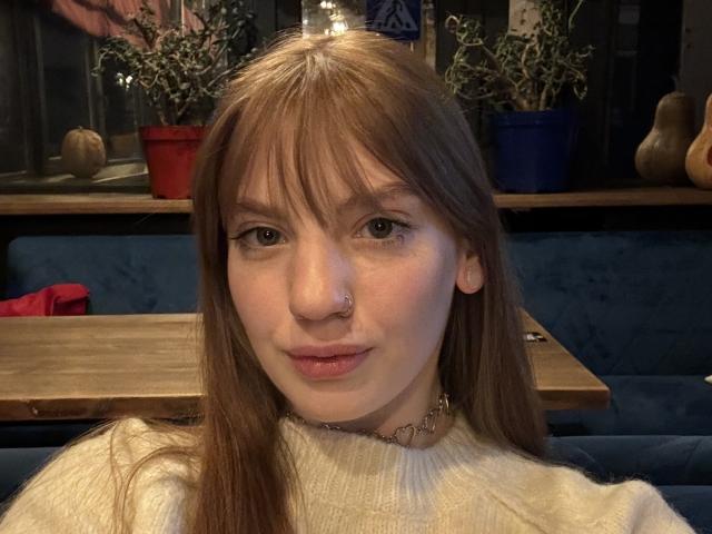 ElsaBj with brown hair, nose ring, and white sweater, sitting in a cozy spot, showing off her cute face and soft lips. Her ass is not visible.