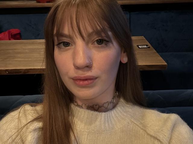 ElsaBj with long brown hair, bangs, and a nose ring, wearing a white sweater and heart-shaped necklace, sitting in a dark blue, wooden-tabled restaurant.