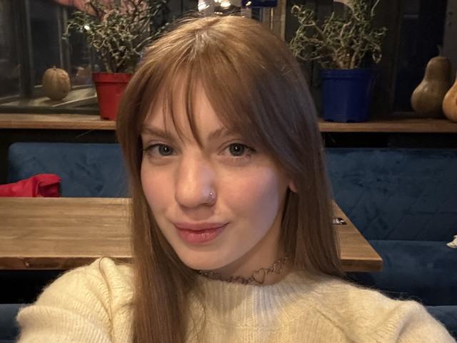 ElsaBj has light brown hair, a nose ring, and a heart-shaped necklace. She's wearing a white sweater and smiling slightly. Her tits are small and she's cute.