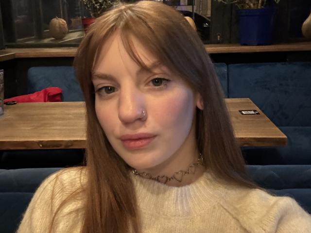 ElsaBj has long brown hair, green eyes, a nose ring, and wears a white sweater with a heart-patterned choker, sitting in a blue booth.