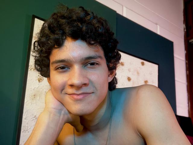 SamuelHills with curly black hair, light brown skin, and a cute smile, shirtless, head resting on hand, wearing a silver necklace, looking at camera.