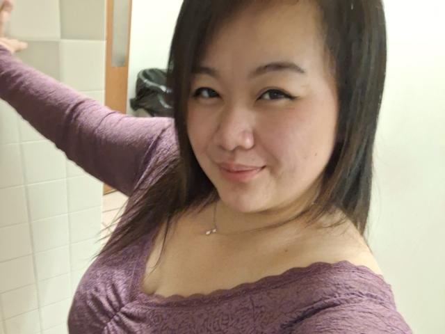 RoxyKay has brown hair, wearing a purple lace top, smiling with a small nose ring, showing her tits slightly, arm raised, in a bathroom.
