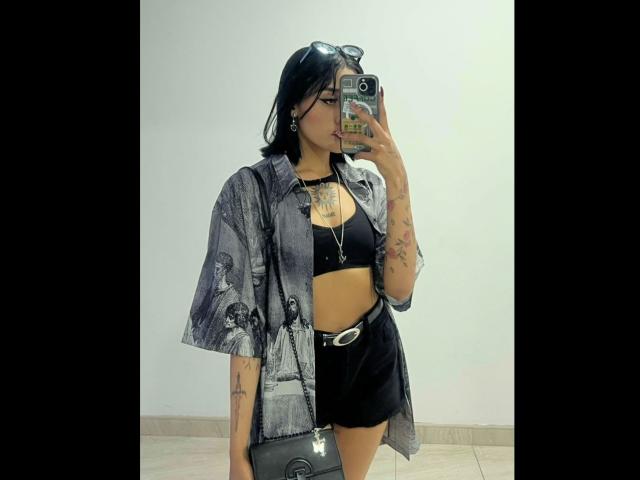 SalomeMille mit schwarzen Haaren, die ein offenes graues Hemd, einen schwarzen Sport-BH, schwarze Shorts und einen Gürtel trägt, hält ein Telefon, zeigt Tattoos, auf einem Selfie im Spiegel..