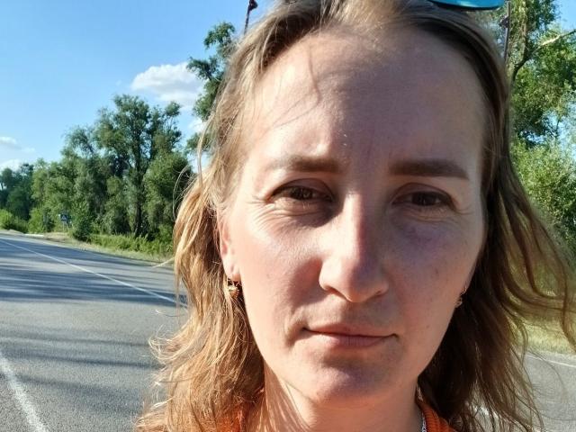 Silfietta's face, showing a slight smile, with light brown hair, small tits, and brown eyes, wearing a orange top, standing on a sunny road with trees in background.
