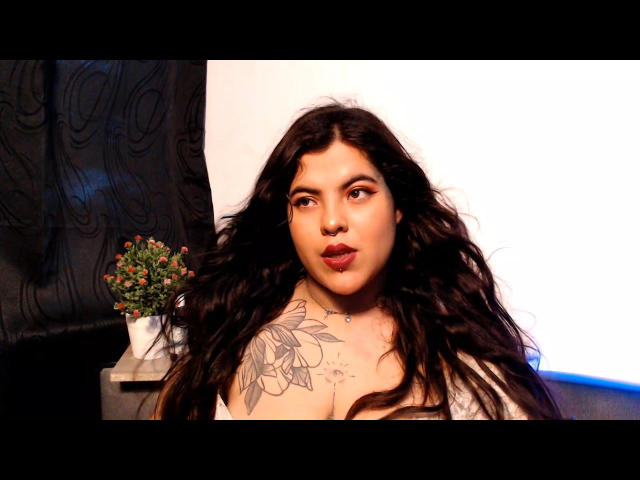 LiianaCooper with long, wavy black hair, pouty lips, and a large floral tattoo on her right shoulder. She's topless, showing her tits, against a white background.