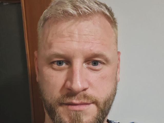 AlexMan69 has a chubby face, blonde hair, blue eyes, and a full beard with a slight smile. His skin is slightly wrinkled and his nose is small.