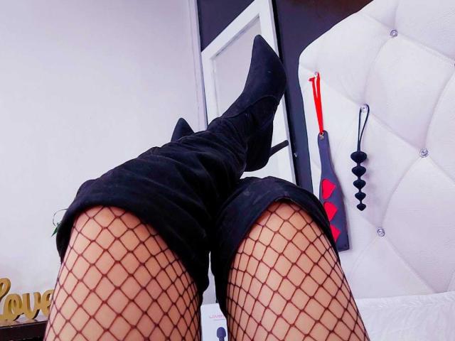 CamilaBardot's legs in black fishnet stockings and black thigh-high boots, showing a black butt plug with red tip on white tufted headboard.