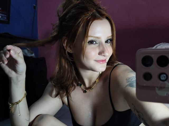 RuivaK with red hair, black tank top, holding phone, showing cleavage, tattoo on arm, gold necklace, and bracelet, smiling, holding hair, pink wall in background.
