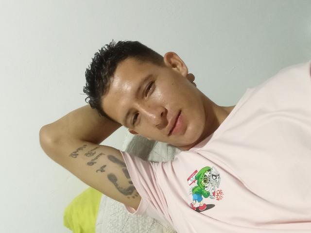 FireLion, with short curly black hair and tattoos on his right arm, wears a pink t-shirt with a green cartoon character, lying down, eyes half-open, looking relaxed.