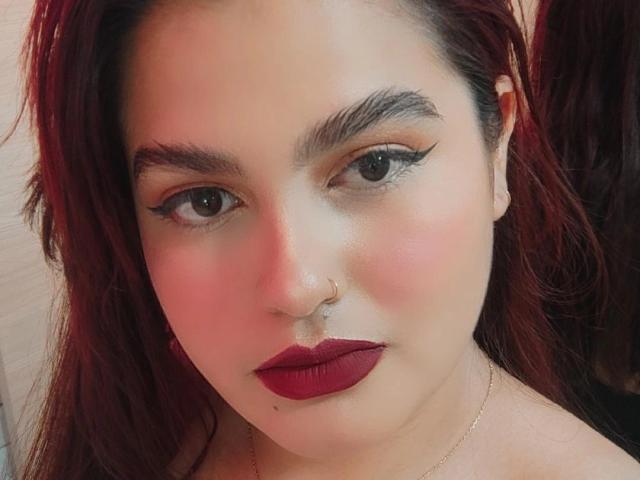 AbbyLester has red lipstick, dark eyeliner, a nose ring, and red hair. She's looking at the camera with a slight blush on her pale skin. Her tits are not visible.