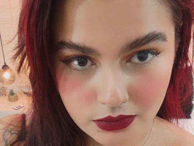 Abby Lester hat rotes Haar, dunkle Augenbrauen, schwarzen Eyeliner, roten Lippenstift, einen Nasenring und ein dezentes Tattoo an ihrer rechten Schulter. Ihr Gesicht ist leicht gerötet..