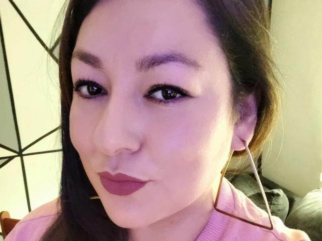 SakotoDika has light skin, purple eyeshadow, and pink lipstick, wearing a pink shirt with white earbuds, and dark brown hair. Her tits are not visible.