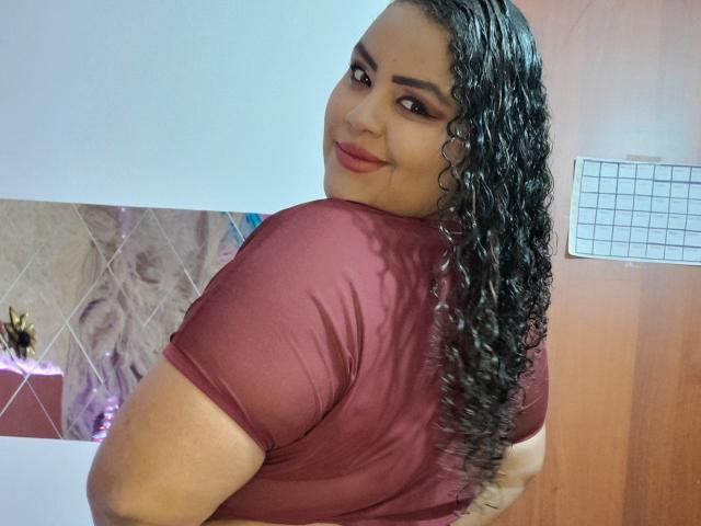 MelannieCarter with curly black hair, brown skin, and red lipstick, wearing a maroon shirt, looking over her shoulder, smiling, showing her back and slightly turned ass.